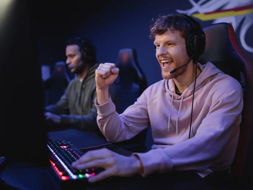 Een jonge esports-speler viert een overwinning achter een gaming-opstelling met een headset en een RGB-toetsenbord, terwijl een geconcentreerde teamgenoot op de achtergrond aan het spelen is